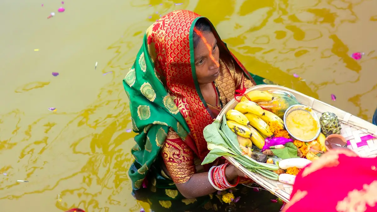 A woman standing in the water holding fruit and diyas as a Chhath offering